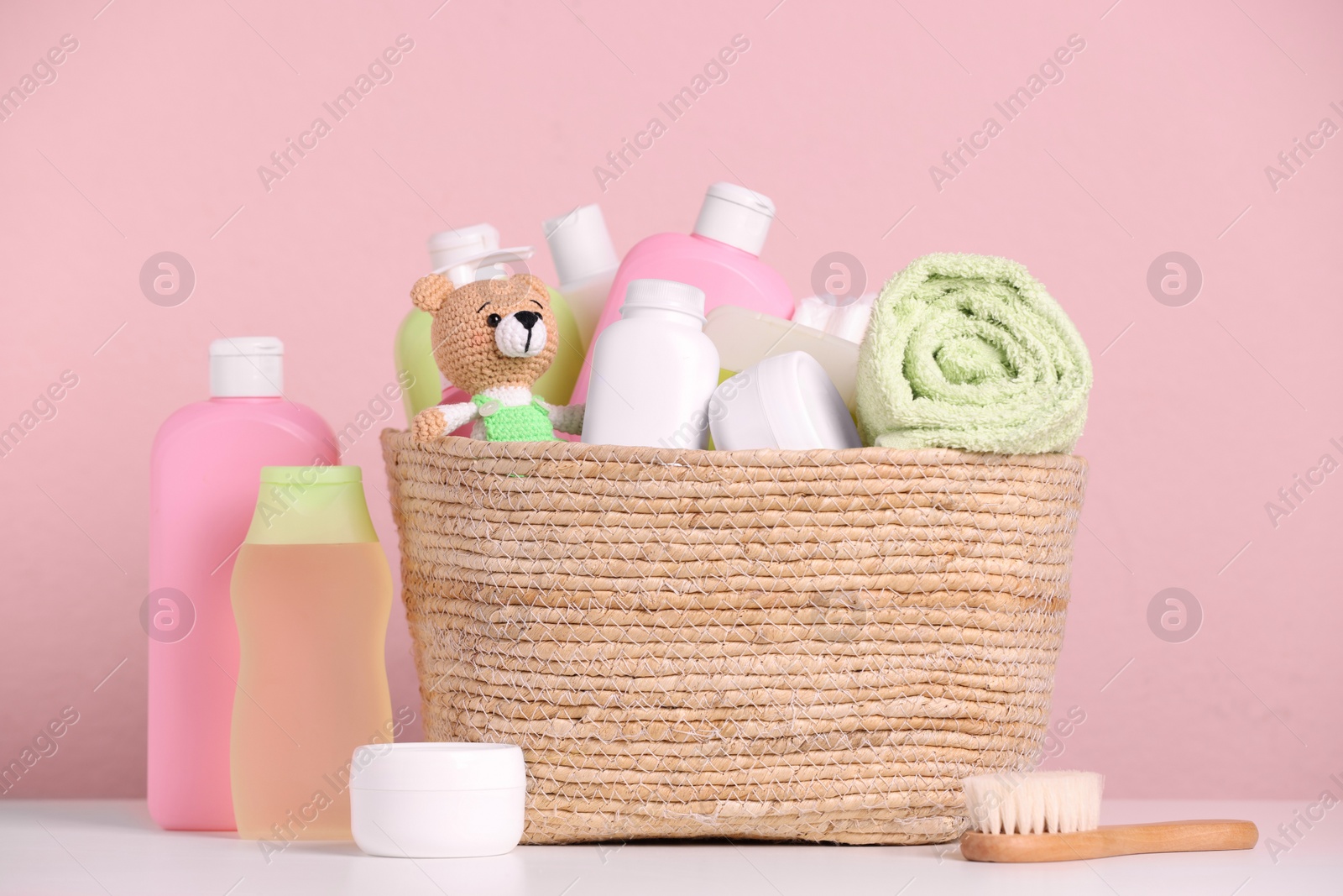 Photo of Baby cosmetic products, bath accessories and toy in wicker basket on white table against pink background