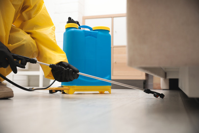 Photo of Pest control worker spraying pesticide under furniture indoors, closeup