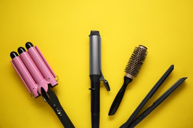 Different curling irons, hair straightener and round brush on yellow background, flat lay