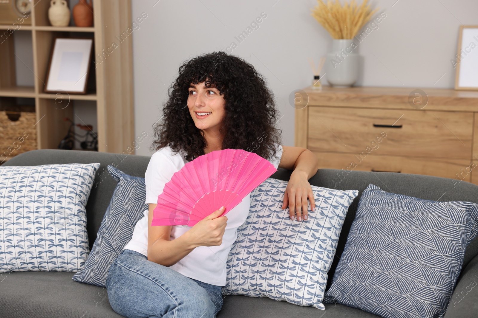 Photo of Young woman waving pink hand fan to cool herself on sofa at home