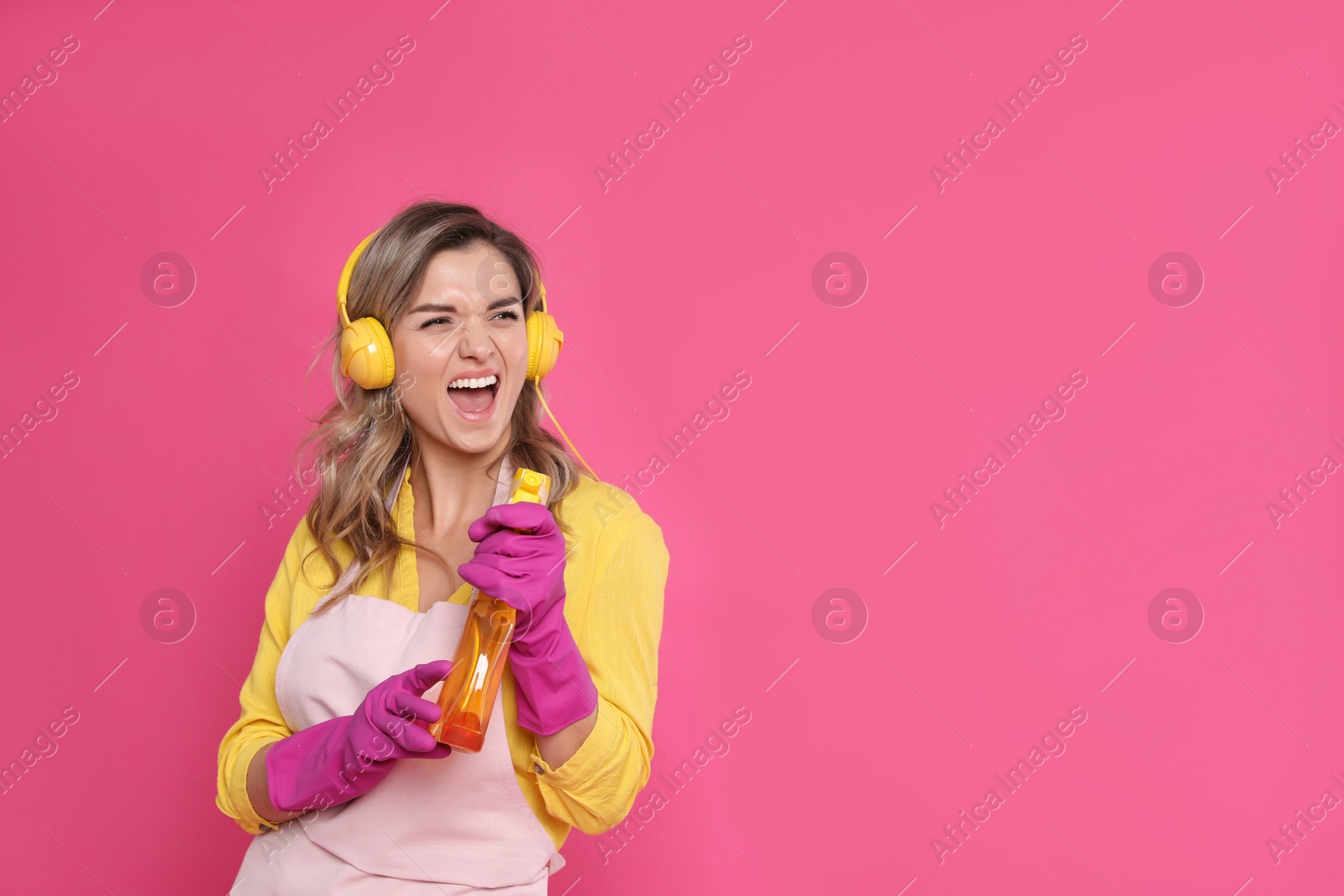 Photo of Beautiful young woman with headphones and bottle of detergent singing on pink background. Space for text