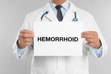 Doctor holding sign with word HEMORRHOID on light grey background, closeup
