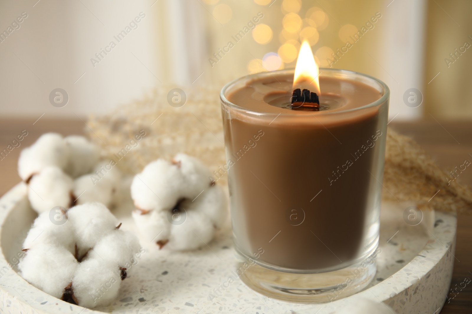 Photo of Burning candle with wooden wick in glass holder near cotton flowers on white tray