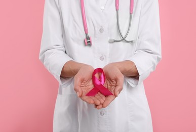 Doctor with stethoscope holding pink ribbon on color background, closeup. Breast cancer awareness