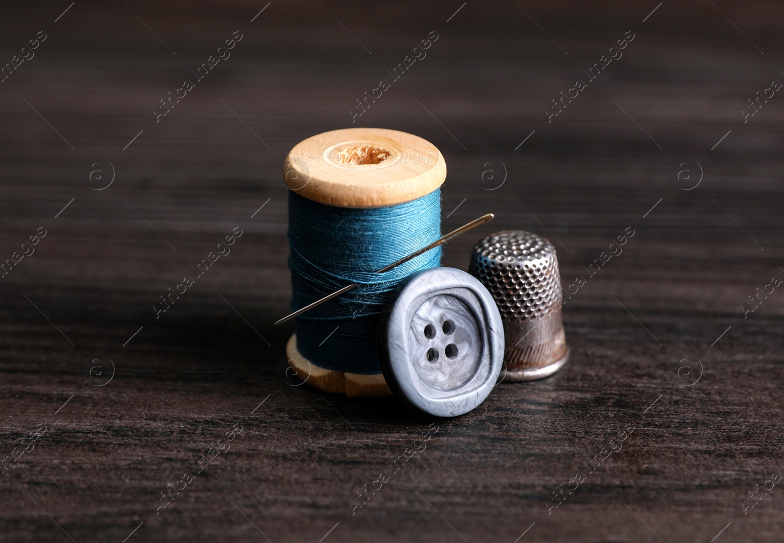 Photo of Spool of light blue thread with needle, button and thimble on wooden table. Sewing accessories