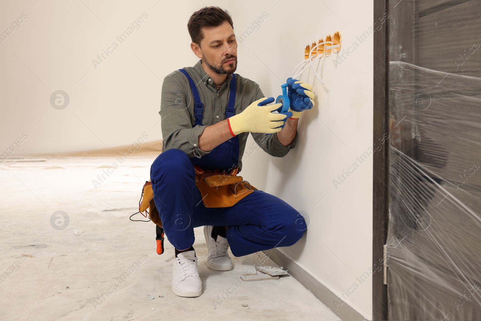 Photo of Electrician in uniform with insulating tape repairing power socket indoors