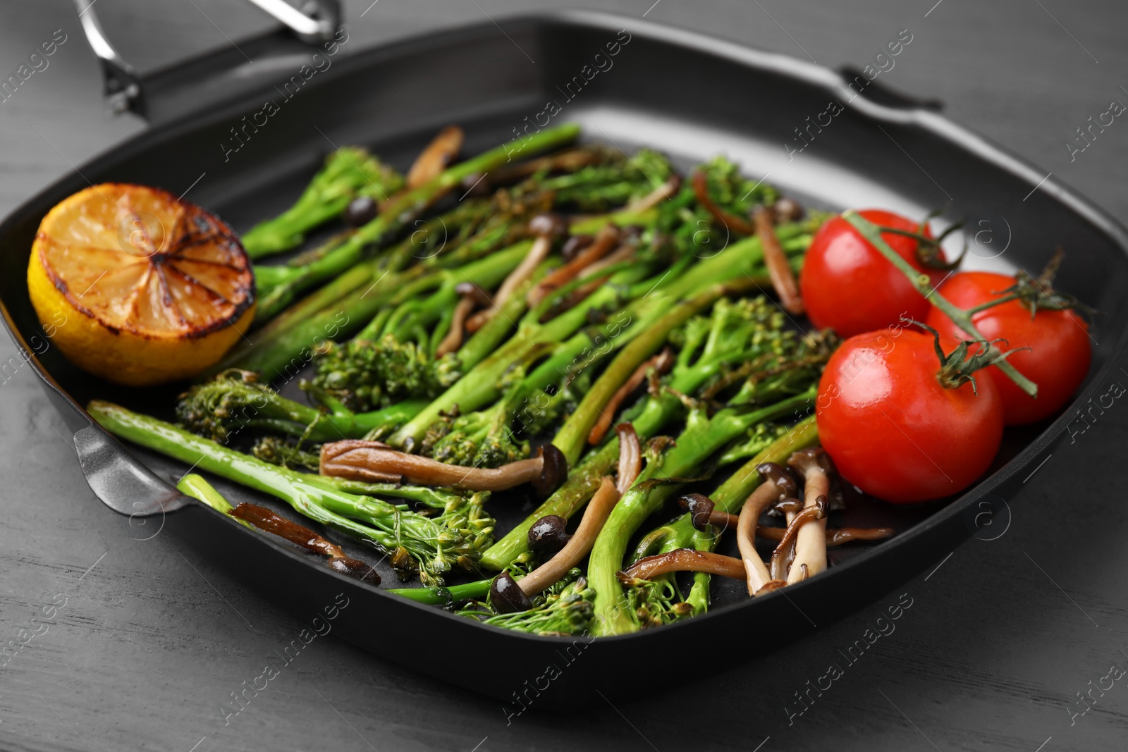 Photo of Grill pan with tasty cooked broccolini, mushrooms, tomatoes and lemon on grey wooden table, closeup