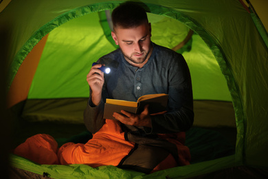 Photo of Young man with flashlight reading book in tent