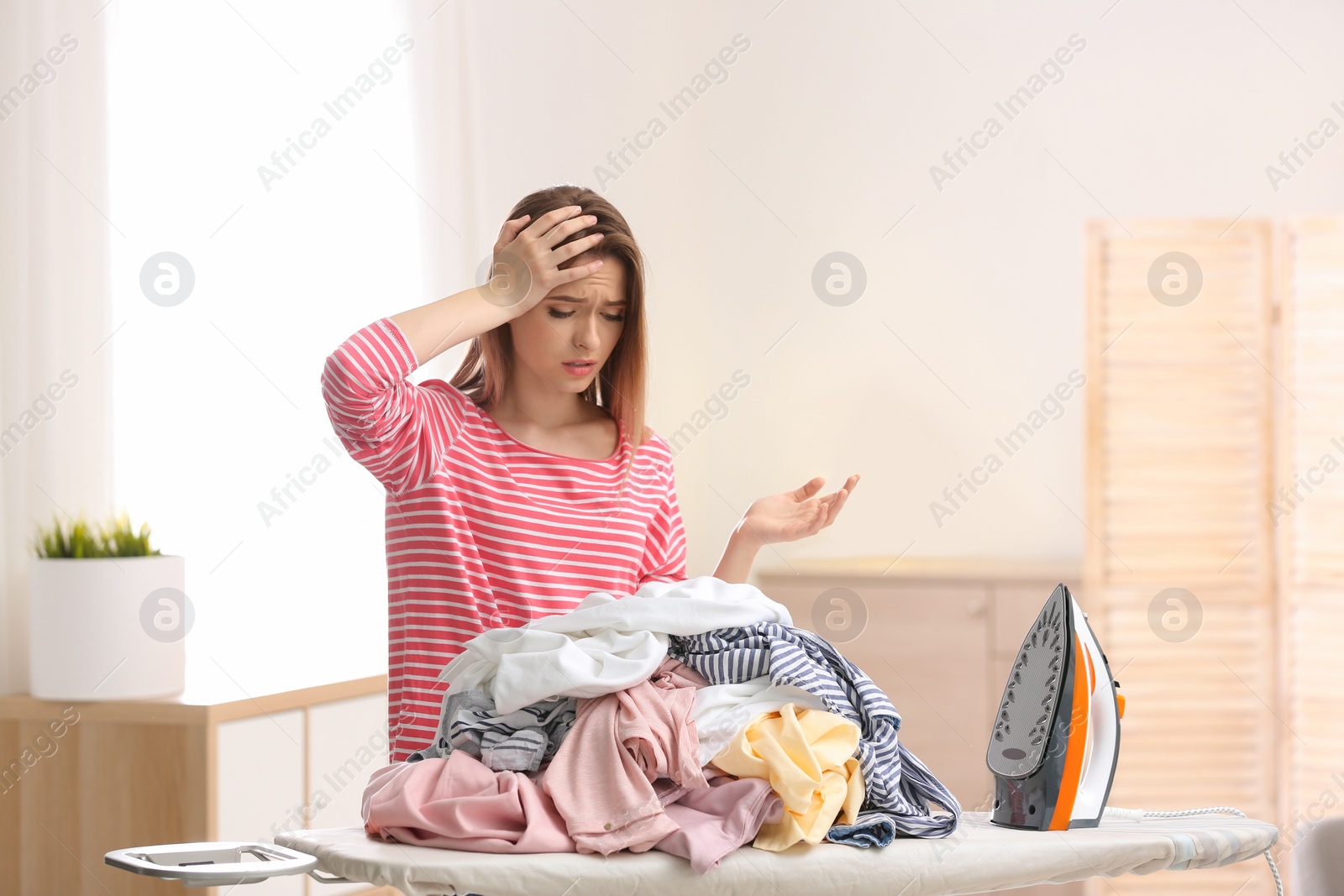 Photo of Young tired woman with clean laundry at ironing board indoors