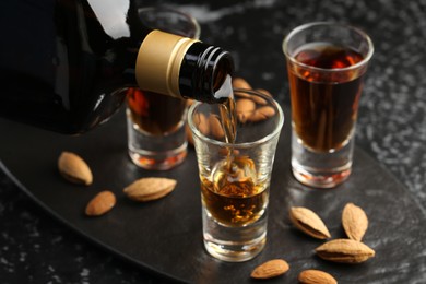 Pouring tasty amaretto liqueur into glass and almonds on black table, closeup