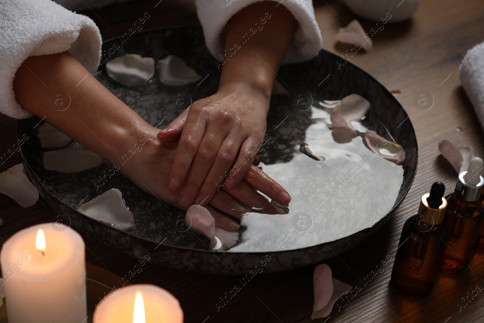 Photo of Woman soaking her hands in bowl of water and flower petals at wooden table, closeup. Spa treatment