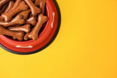 Red bowl with bone shaped dog cookies on yellow background, top view. Space for text
