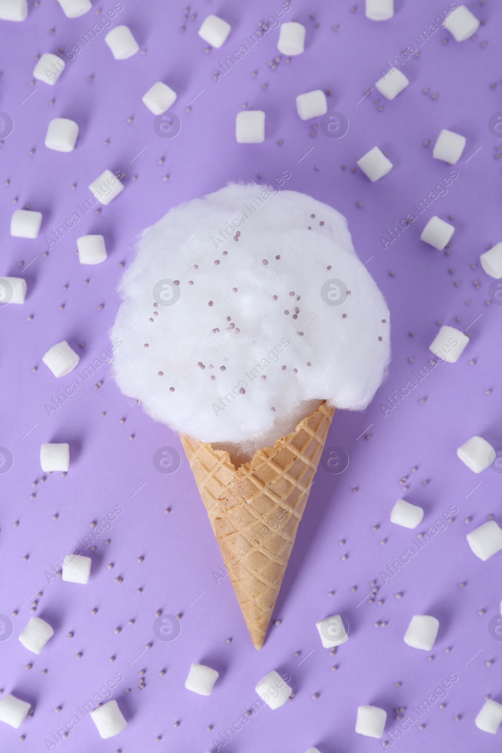 Photo of Sweet cotton candy in waffle cone surrounded by marshmallows on purple background, flat lay