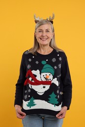 Happy senior woman in deer headband showing Christmas sweater on orange background