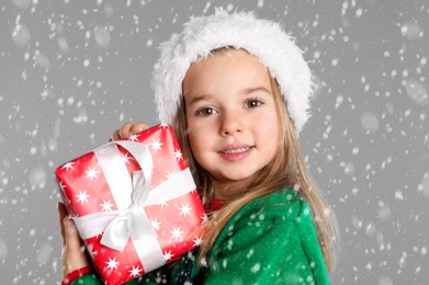 Image of Cute child in Santa hat with Christmas gift under snowfall on grey background