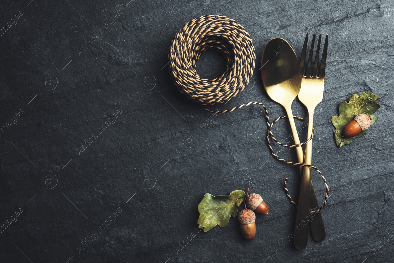 Photo of Cutlery, acorns and rope on black slate background, flat lay with space for text. Table setting elements