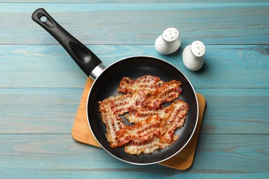 Delicious bacon slices in frying pan on blue wooden table, flat lay