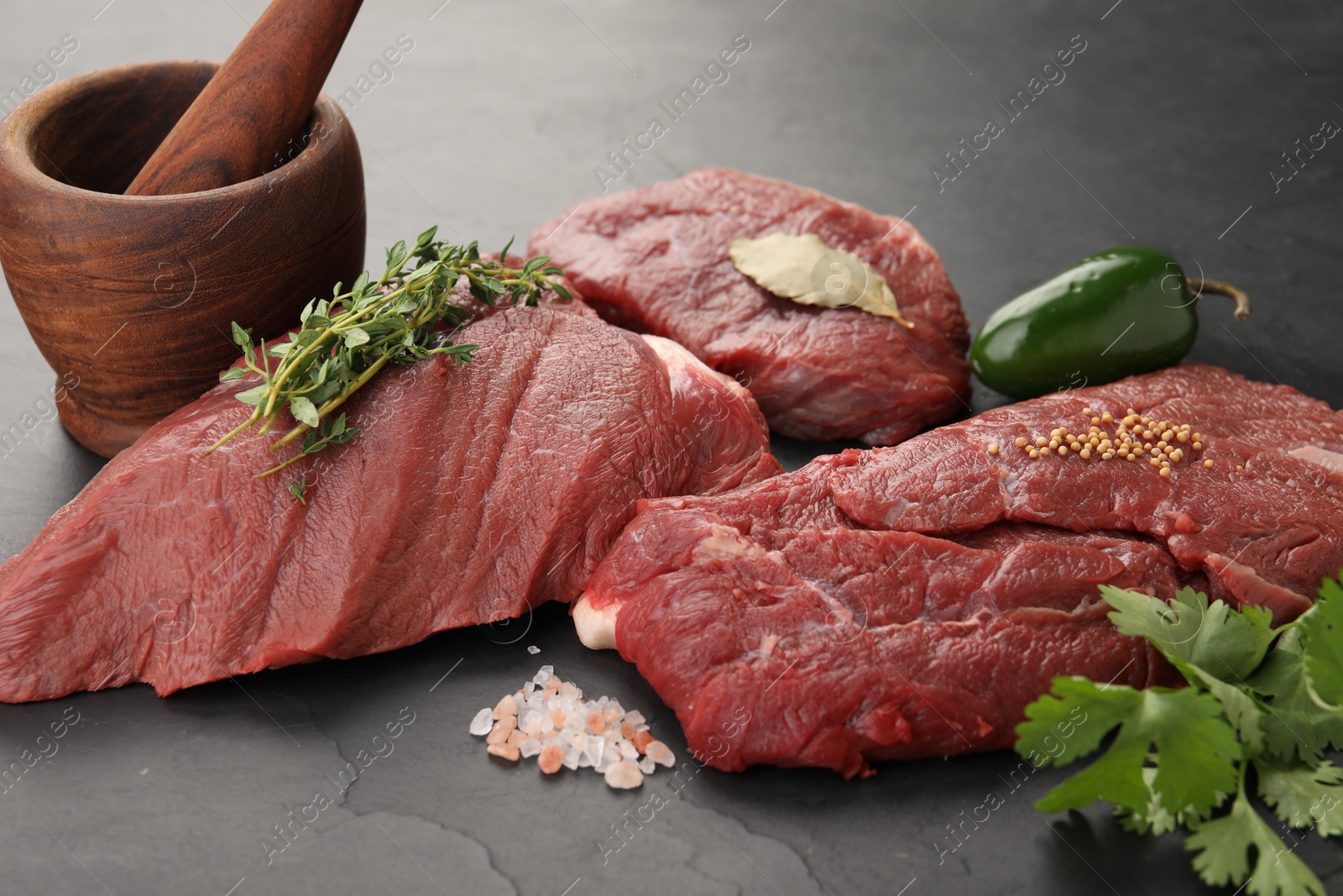 Photo of Pieces of raw beef meat, mortar with pestle and spices on black table, closeup