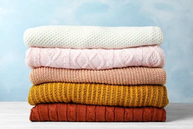 Photo of Stack of warm clothes on white wooden table against light blue background. Autumn season