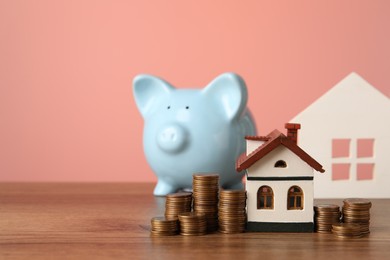 Photo of House models, piggy bank and stacked coins on wooden table against pink background, selective focus. Space for text