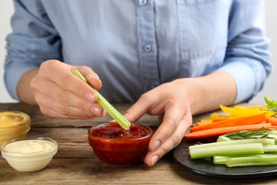 Woman dipping celery stick in sauce at wooden table, closeup