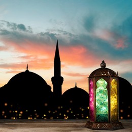 Decorative Arabic lantern on wooden surface and silhouette of mosque at sunset on background, space for text