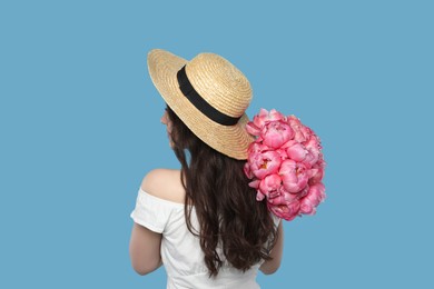 Photo of Beautiful young woman in straw hat with bouquet of pink peonies against light blue background