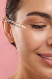 Photo of Beautiful young woman applying serum onto her face on pink background, closeup