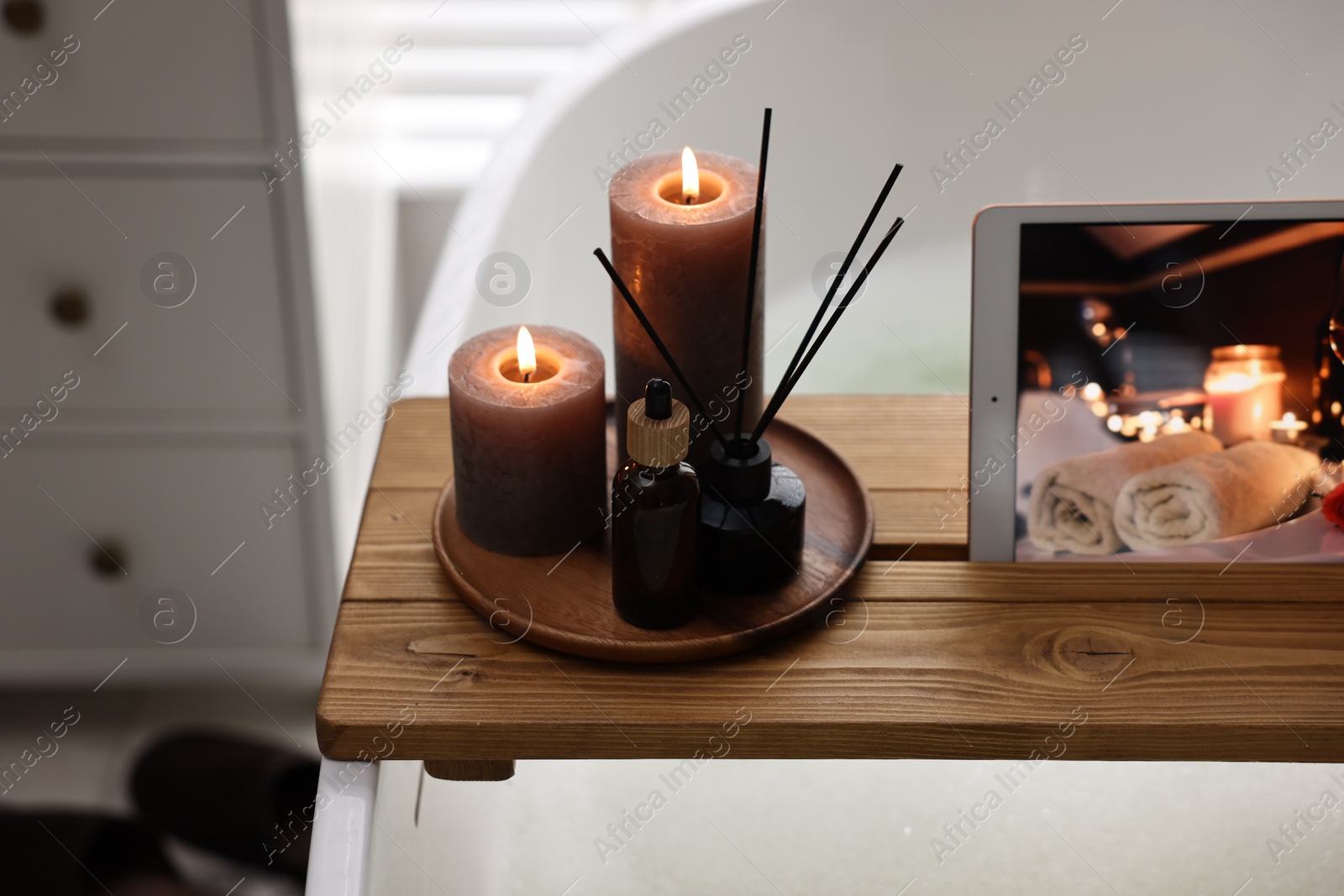 Photo of Wooden tray with tablet, burning candles and aroma products on bathtub in bathroom