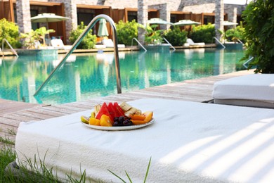 Photo of Plate with fresh fruits on sun lounger near outdoor swimming pool. Luxury resort