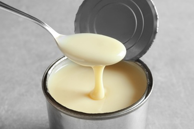 Photo of Condensed milk pouring from spoon into tin can on grey background, closeup. Dairy product