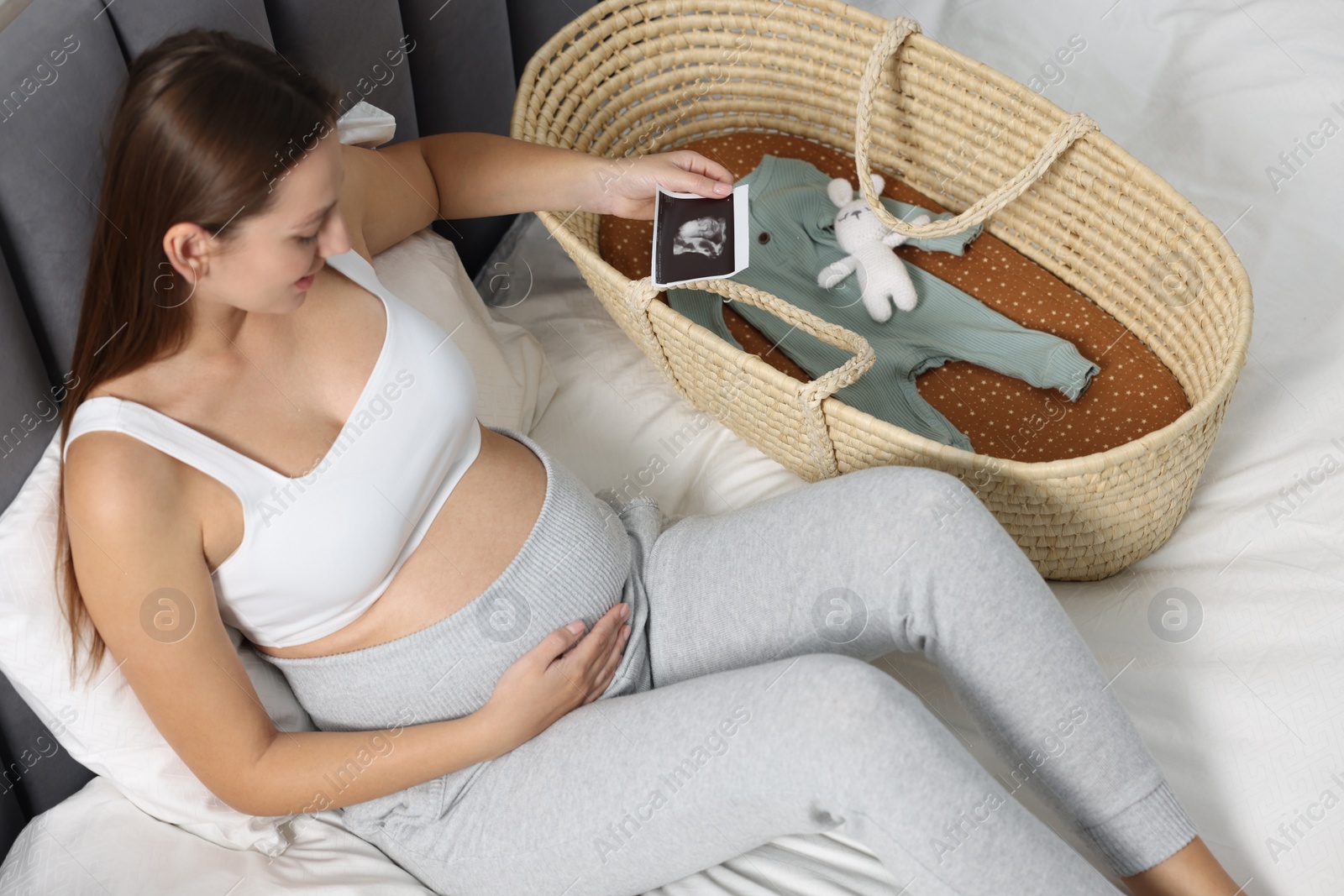 Photo of Pregnant woman with ultrasound picture of baby and basket on bed, above view