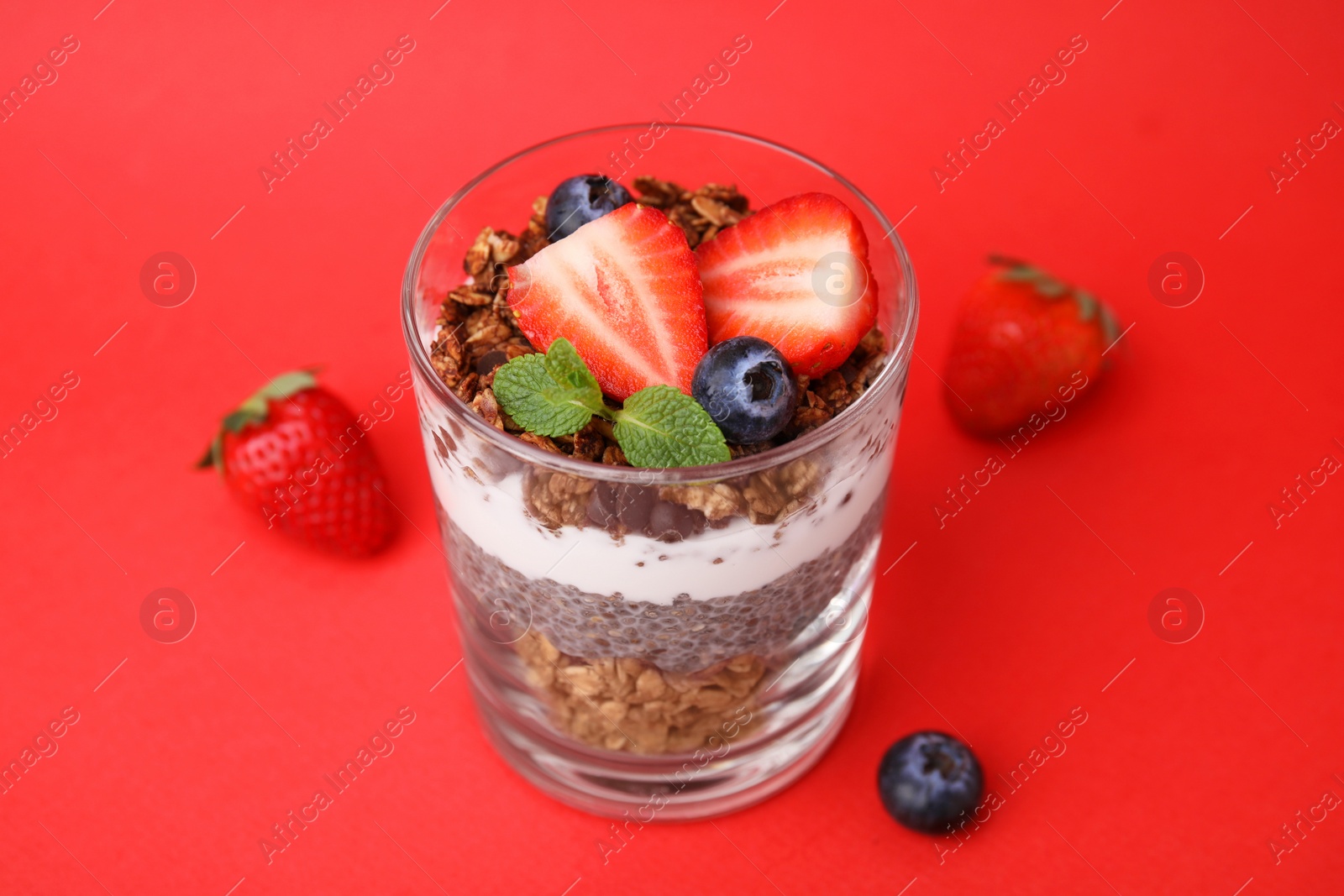 Photo of Tasty granola with berries, yogurt and chia seeds in glass on red background, closeup