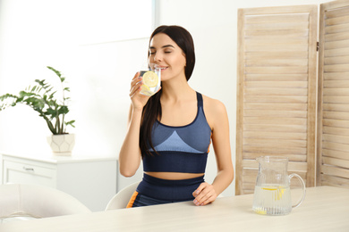 Photo of Beautiful sportive woman drinking lemon water at table indoors