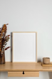 Photo of Empty photo frame, cup and vase with dry decorative leaves on wooden table. Mockup for design