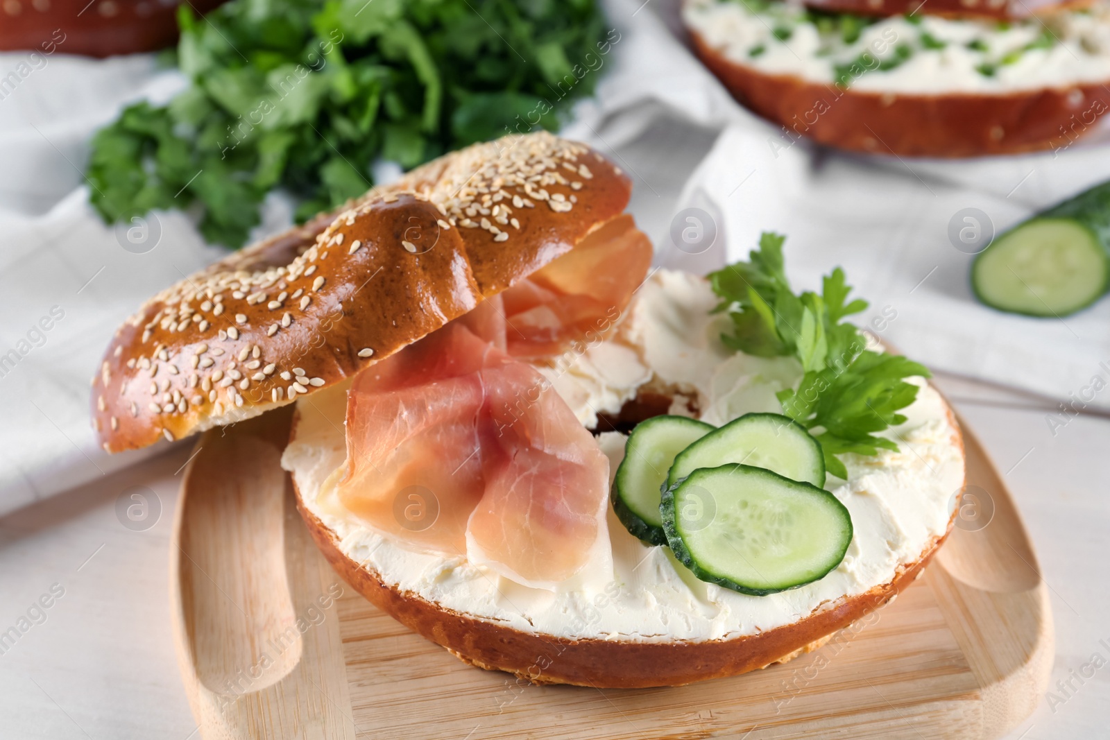 Photo of Delicious bagel with cream cheese, jamon, cucumber and parsley on wooden board, closeup