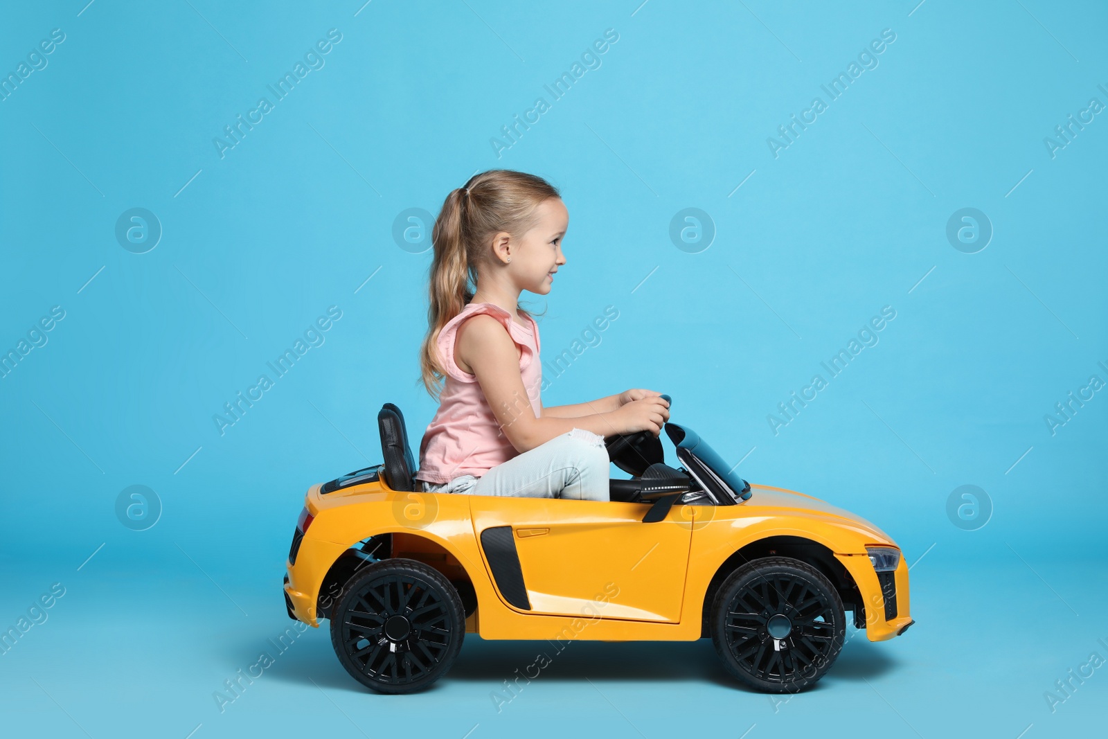 Photo of Cute little girl driving children's electric toy car on light blue background