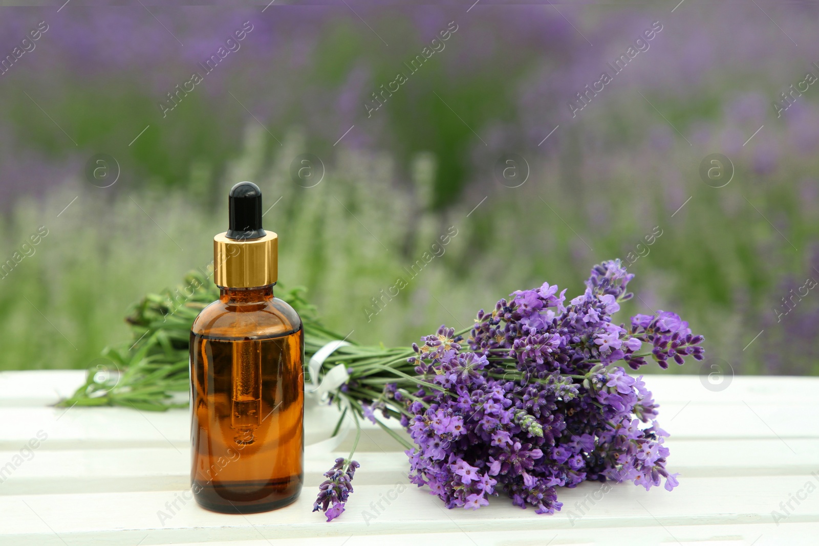 Photo of Bottle of essential oil and lavender flowers on white wooden table in field