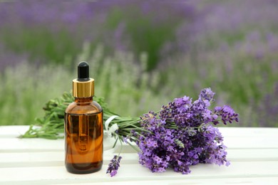 Bottle of essential oil and lavender flowers on white wooden table in field