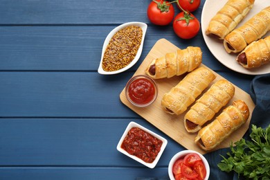 Photo of Delicious sausage rolls and ingredients on blue wooden table, flat lay. Space for text