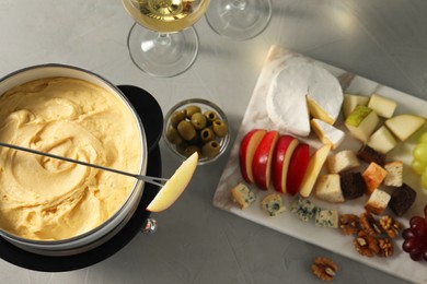 Fork with piece of apple, melted cheese in fondue pot, wine and snacks on grey table, flat lay