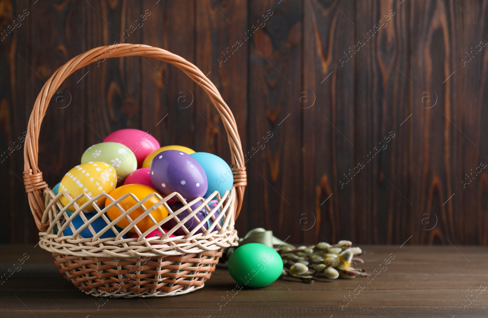 Photo of Colorful Easter eggs in wicker basket and willow branches on wooden background. Space for text