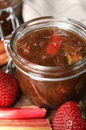 Photo of Jar of tasty rhubarb jam, fresh stems and strawberries on wooden table, closeup