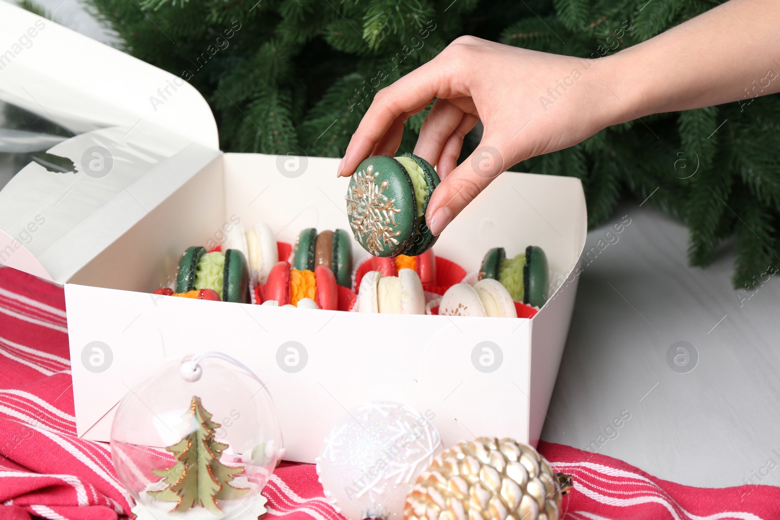 Photo of Woman with box of decorated Christmas macarons at white table, closeup