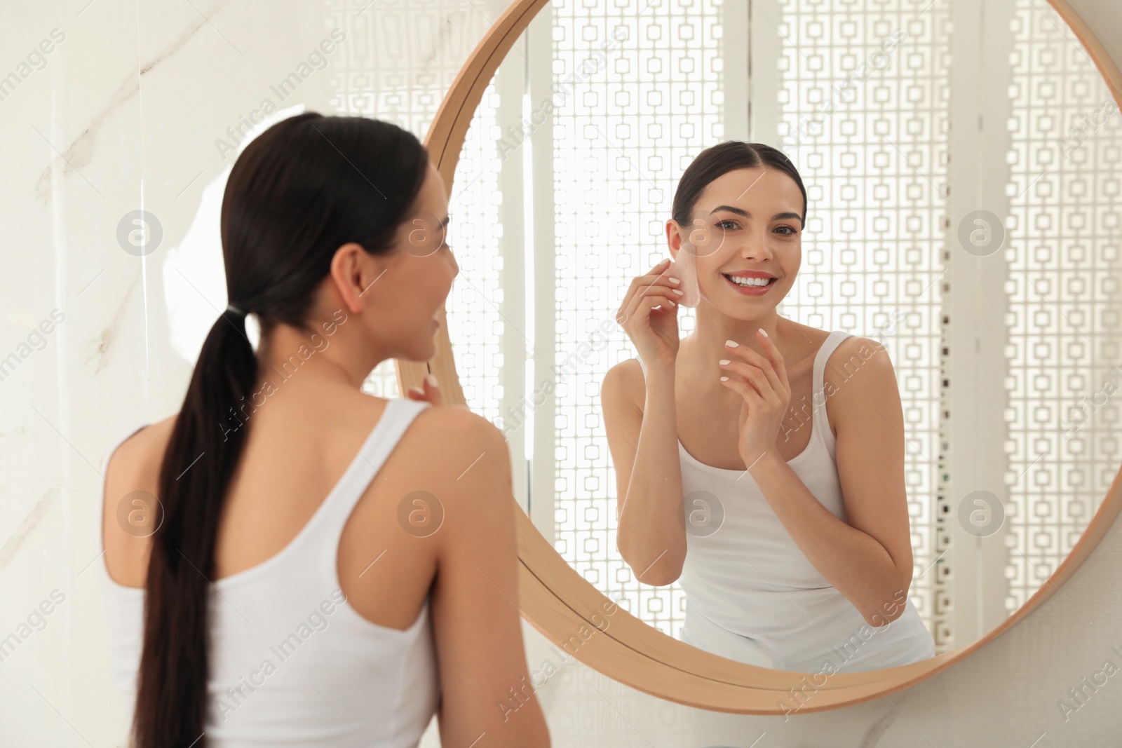 Photo of Beautiful young woman doing facial massage with gua sha tool in front of mirror at home