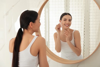Photo of Beautiful young woman doing facial massage with gua sha tool in front of mirror at home