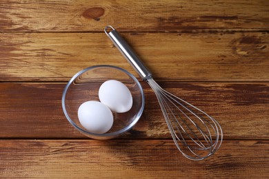 Photo of Metal whisk and eggs in bowl on wooden table, flat lay