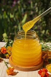 Photo of Taking delicious fresh honey with dipper from glass jar surrounded by beautiful flowers on table in garden