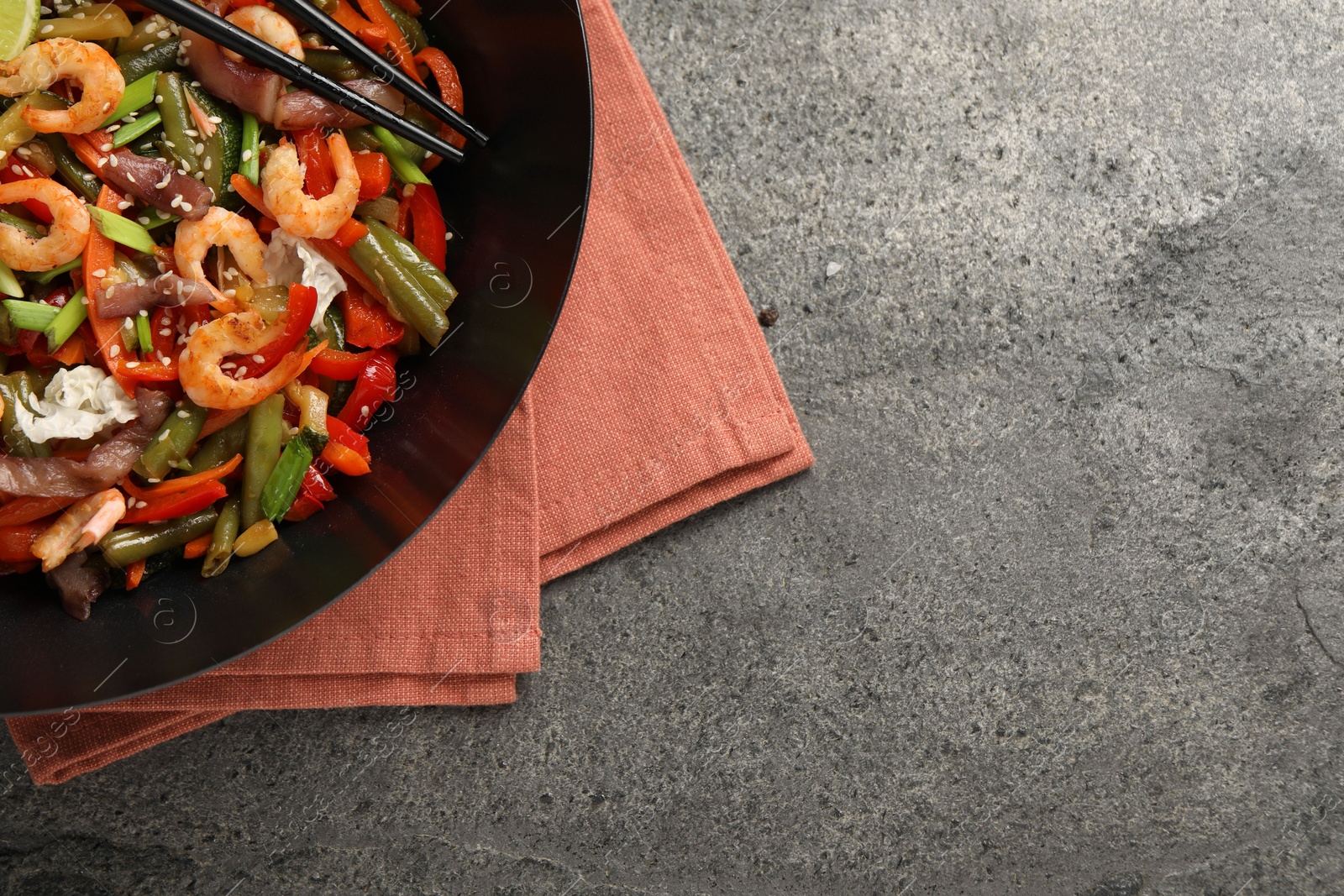 Photo of Shrimp stir fry with vegetables in wok and chopsticks on grey table, top view. Space for text