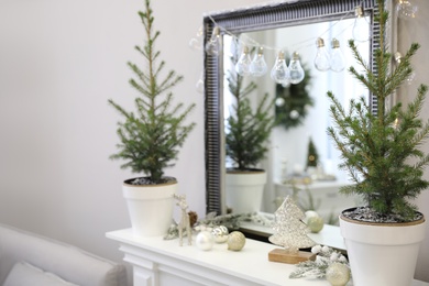 Photo of Little fir trees and Christmas decorations on shelf near mirror in room. Stylish interior design